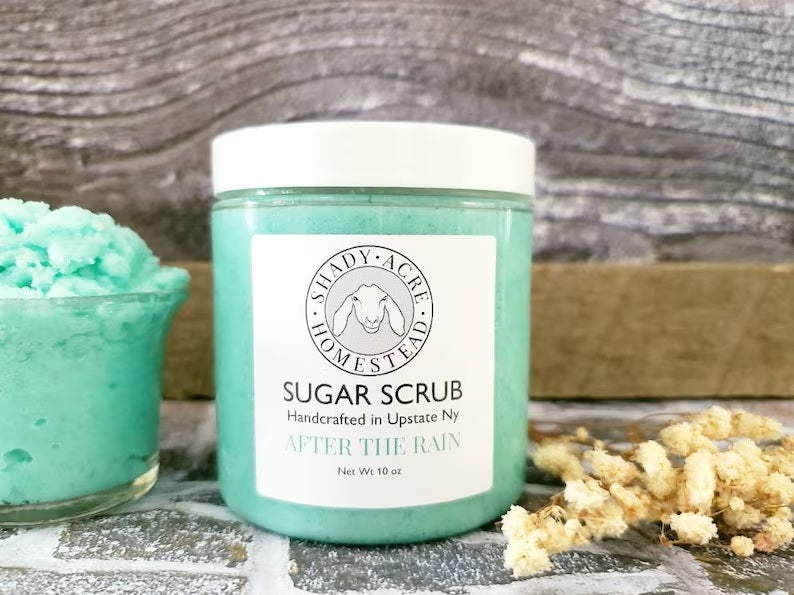 Jar of handmade sugar scrub labeled 'After the Rain' on a rustic surface with decorative elements.