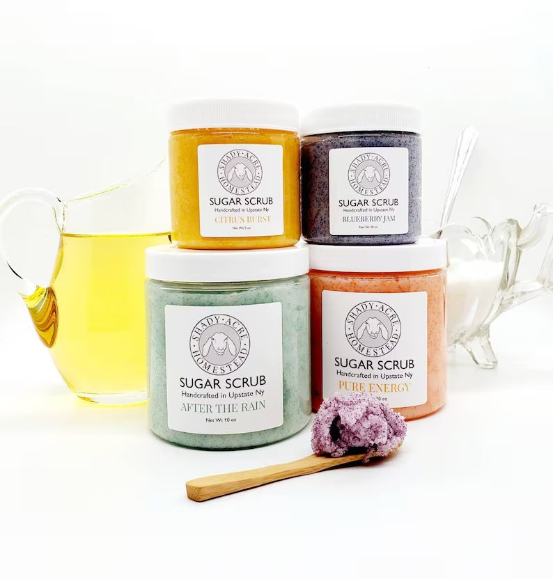 Four jars of handmade sugar scrubs with different scents next to a pitcher of liquid and a wooden spoon.