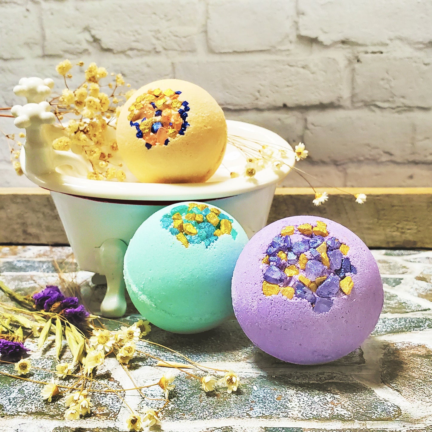 Three decorative bath bombs in pastel colors placed on a rustic surface, with a miniature white bathtub and dried flowers in the background.