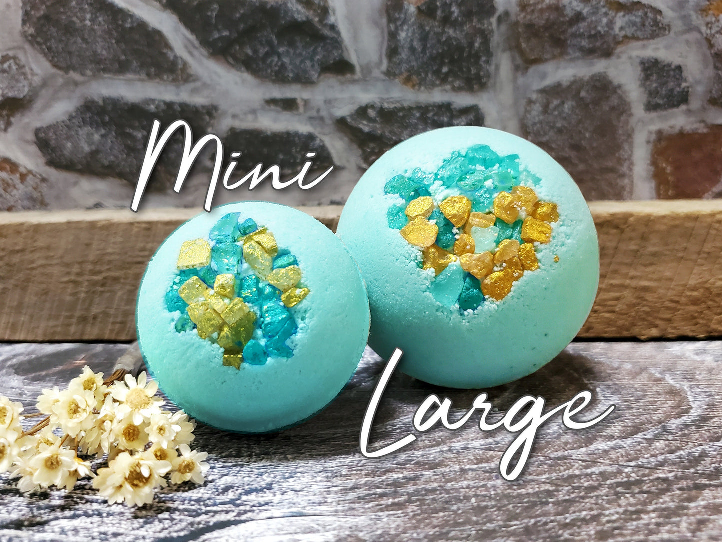 Two blue bath bombs with turquoise and gold glitter, one labeled 'Mini' and the other 'Large', with white flowers in the foreground.