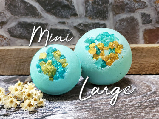 Two blue bath bombs with turquoise and gold glitter, one labeled 'Mini' and the other 'Large', with white flowers in the foreground.