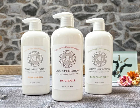 Three bottles of Shady Acre Homestead goat's milk lotion with labels reading 'Pure Energy,' 'Patchouli,' and 'Rosemary Mint' on a wooden surface with a stone background.